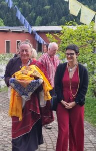 Lama souriant portant des khatas et une pratiquante en tenue bordeaux dans la cour extérieure de Nigu Karma Ling, avec drapeaux de prières et bâtiments du campus en arrière-plan